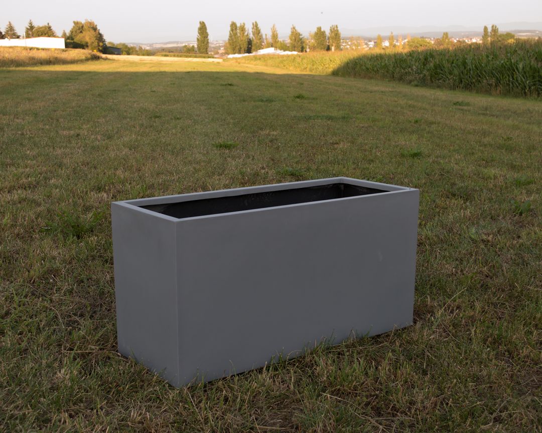 Grauer rechteckiger Pflanzkübel aus Fiberglas ohne Bepflanzung auf einer Wiese vor Feldern und Bäumen im Abendlicht