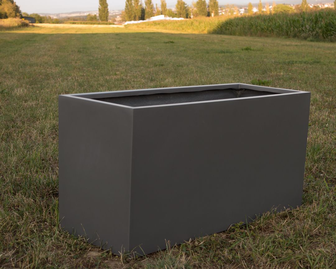 Rechteckiger Pflanzkübel aus Fiberglas in Anthrazit metallic, leer auf einer Wiese mit Feldern im Hintergrund