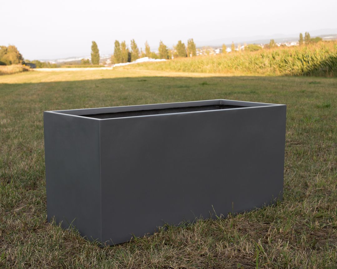 Rechteckiger Pflanzkübel aus Fiberglas in Anthrazit metallic, leer auf einer Wiese vor Feldern und Bäumen im Abendlicht