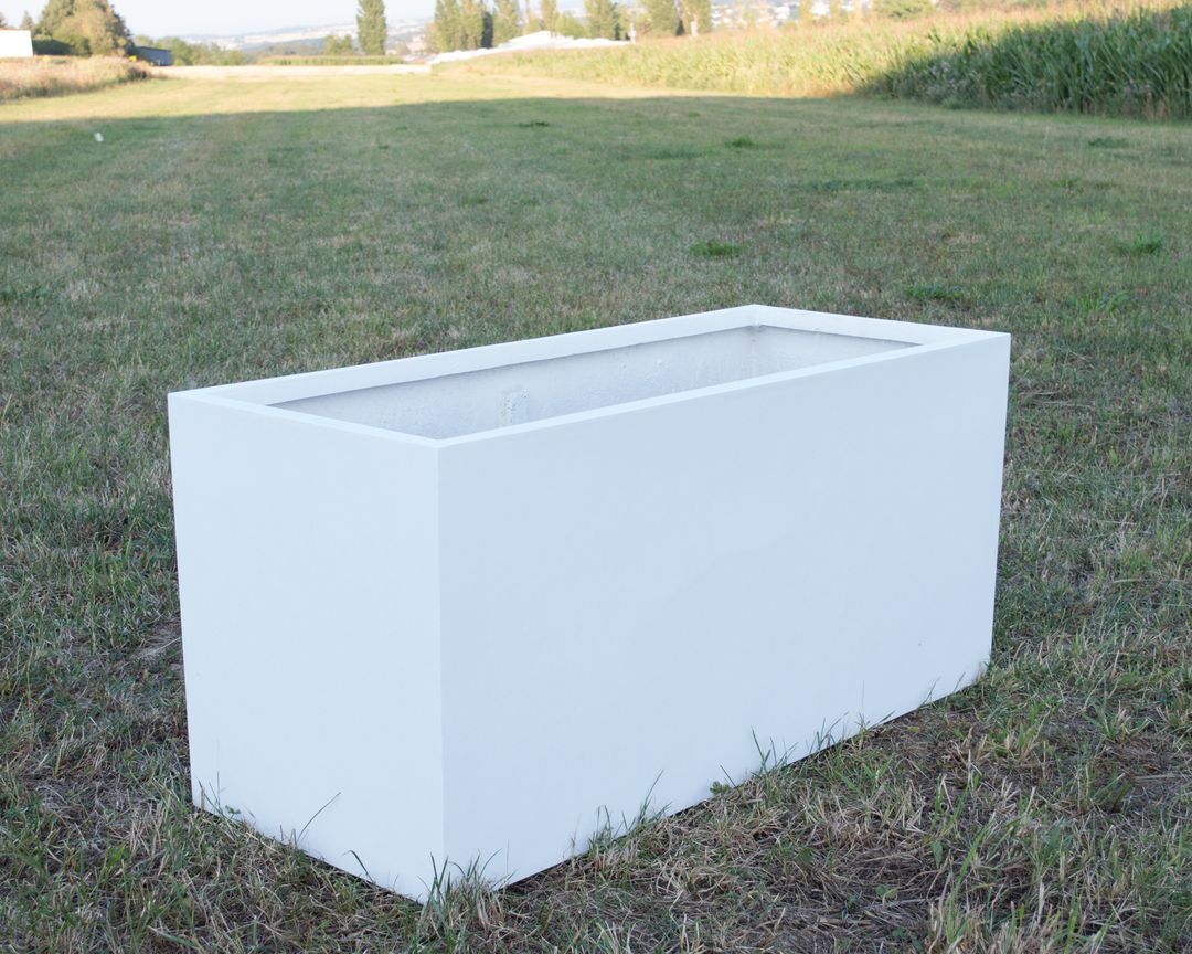 Rechteckiger Pflanzkübel aus Fiberglas in Perlmuttweiß, leer auf einer Wiese vor Feldern und Bäumen