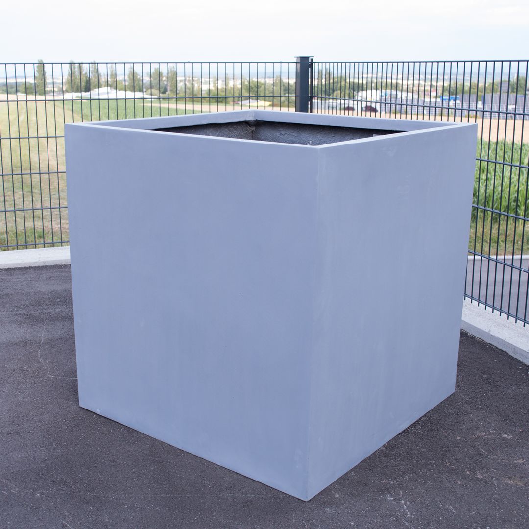 Quadratischer grauer Pflanzkübel aus Fiberglas ohne Bepflanzung auf einer Asphaltfläche vor einem Gitterzaun
