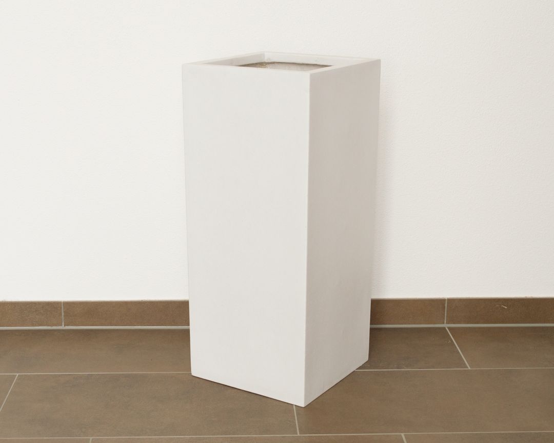 Hohe quadratische Pflanzkübel-Säule aus Fiberglas in Perlmuttweiß vor weißer Wand auf braunem Fliesenboden
