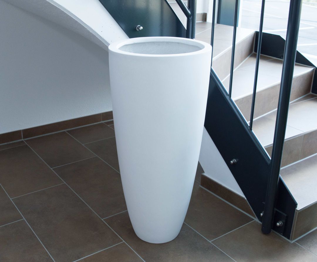 Perlmuttweißer runder konischer Pflanzkübel aus Fiberglas ohne Bepflanzung neben einer Treppe auf braunen Fliesen