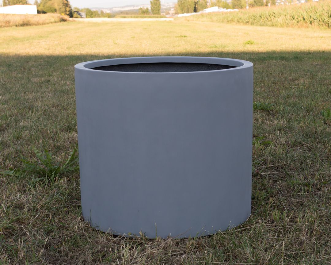 Grauer zylindrischer Pflanzkübel aus Fiberglas ohne Bepflanzung auf einer Wiese vor Feldern und Bäumen