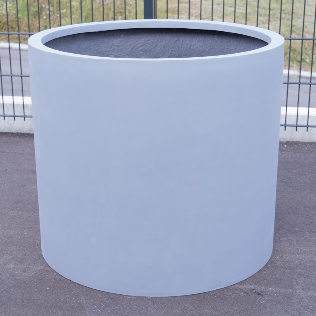 Grauer zylindrischer Pflanzkübel aus Fiberglas ohne Bepflanzung auf Asphalt vor einem Metallgitterzaun