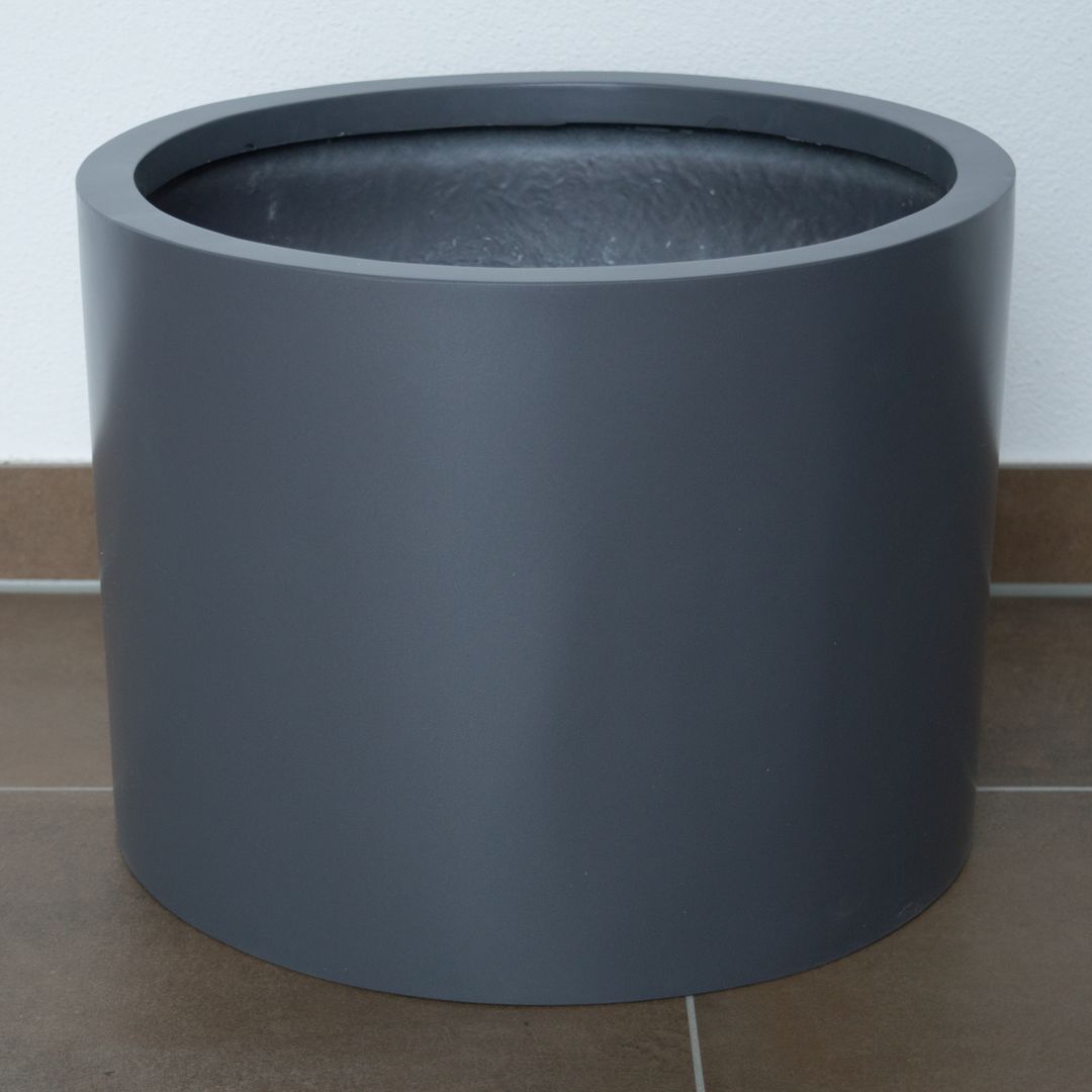 Zylindrischer Pflanzkübel aus Fiberglas in Anthrazit-Metallic ohne Bepflanzung auf braunem Fliesenboden vor weißer Wand