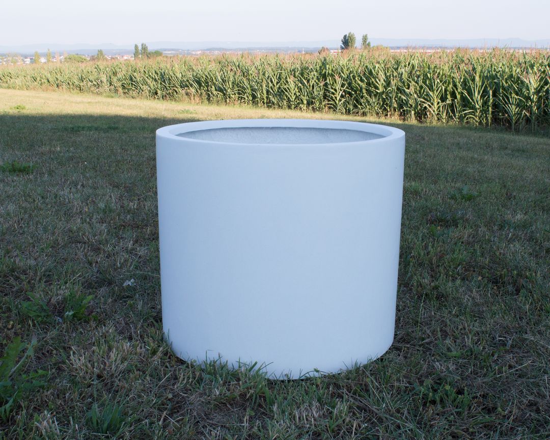 Zylindrischer Pflanzkübel aus Fiberglas in Perlmuttweiß ohne Bepflanzung auf einer Wiese vor einem Maisfeld