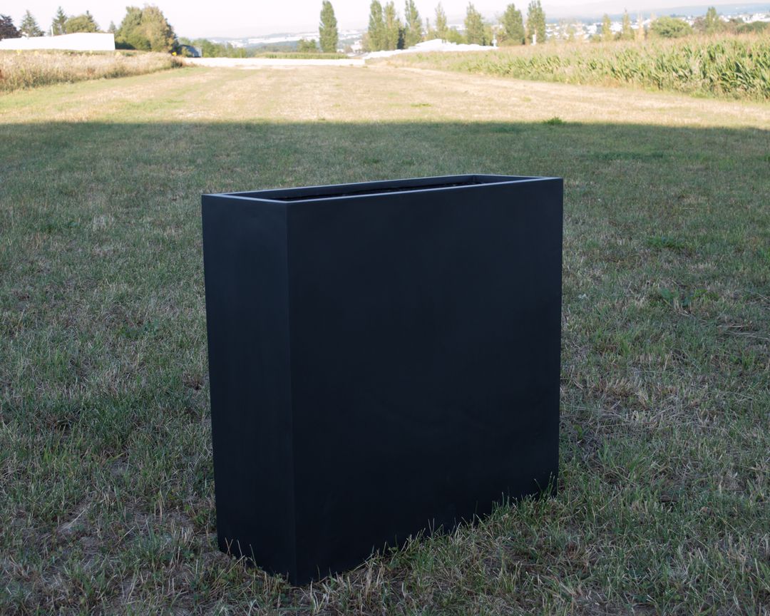 Rechteckiger Pflanzkübel aus Fiberglas in Schwarz-matt ohne Bepflanzung auf einer Wiese vor Feldern und Bäumen