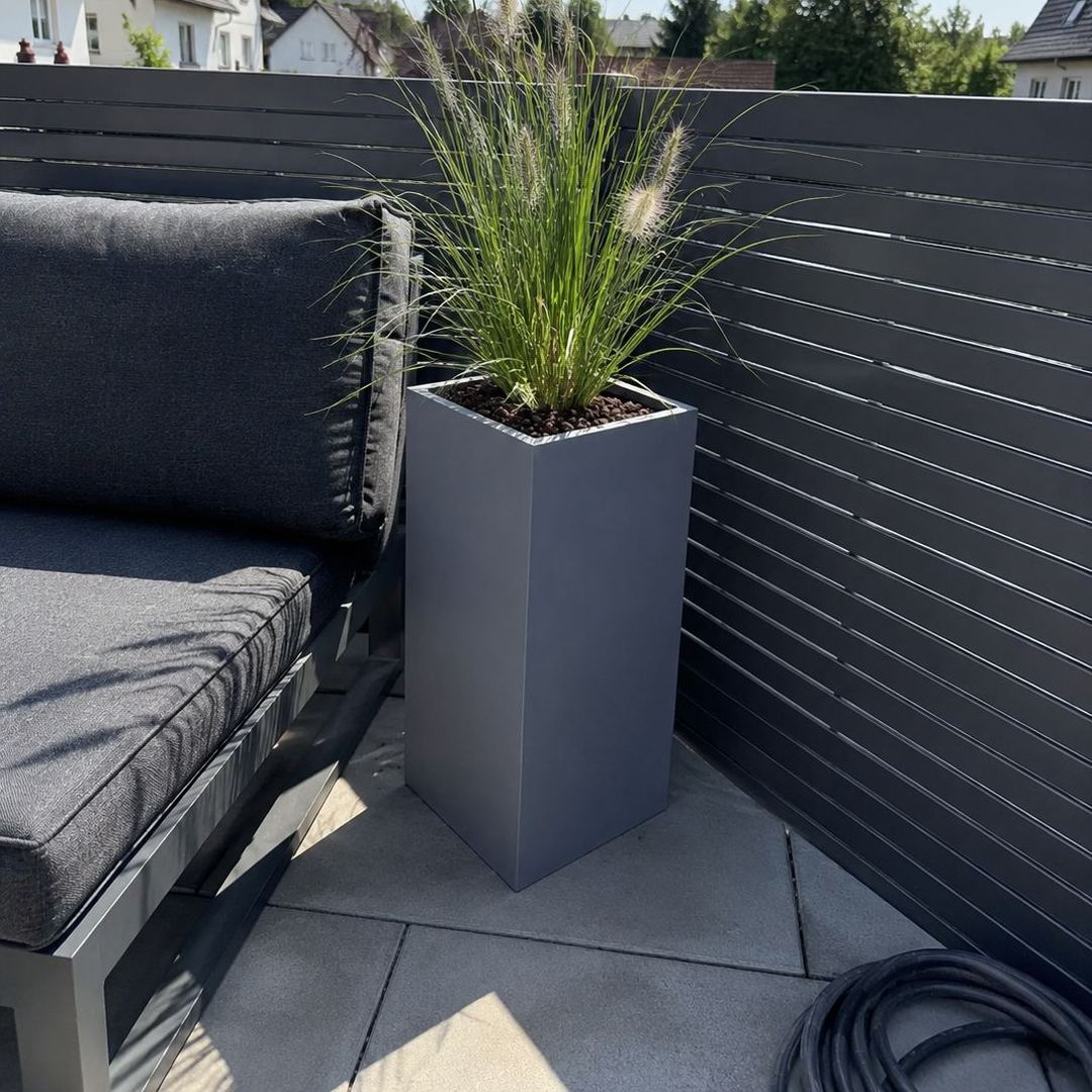 Grauer Pflanzkübel aus Fiberglas in Säulenform mit Lampenputzergras auf Terrasse neben Loungesofa und Sichtschutzzaun