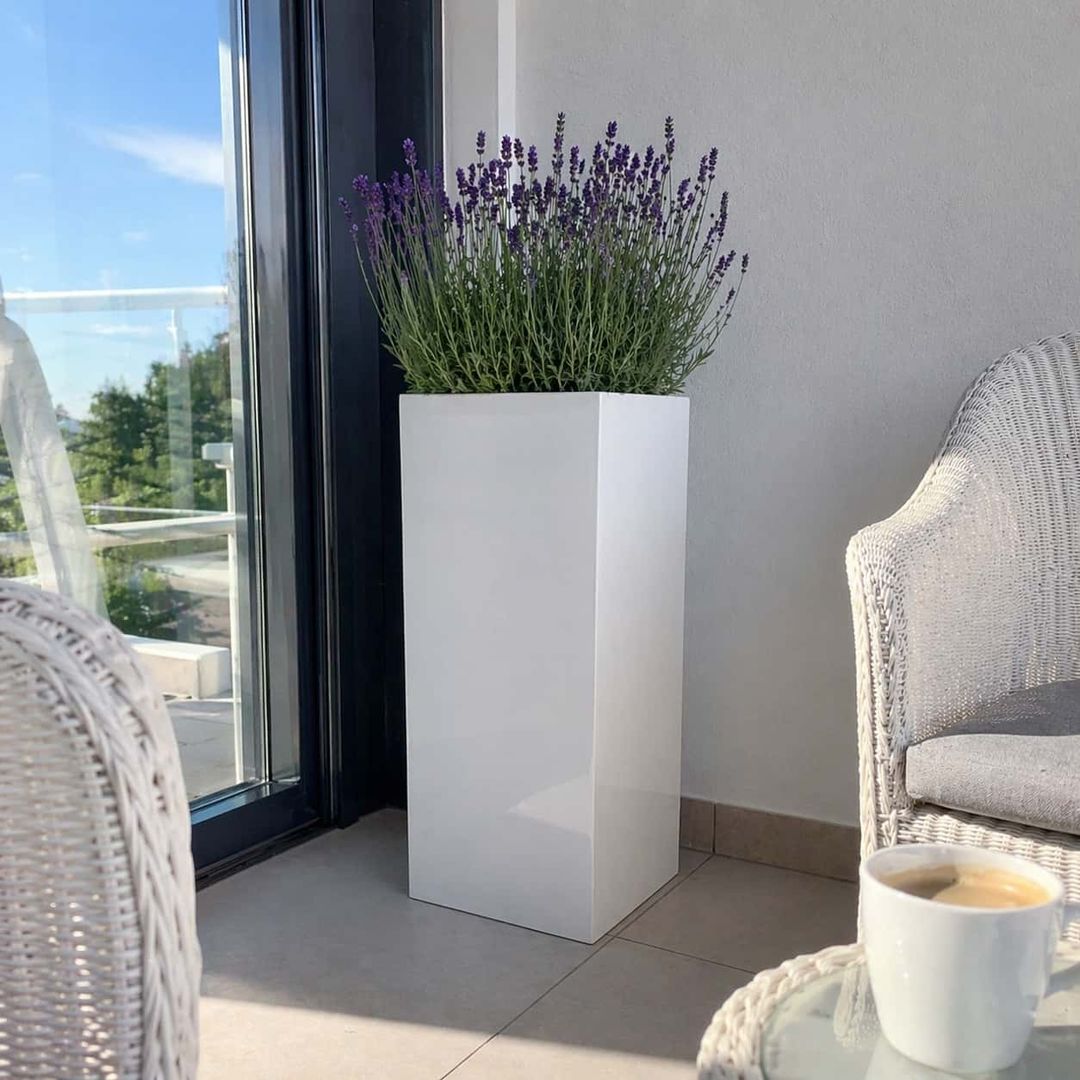 KI-erstellte Abbildung: Hoher Säulen-Pflanzkübel in Hochglanz Weiß mit Lavendel auf einem Balkon neben Rattansesseln und Kaffeetasse