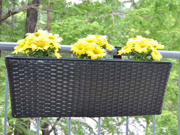 Rechteckiger Pflanzkübel aus Polyrattan in Coffee Braun mit gelben Blüten am Balkongeländer vor grünen Bäumen
