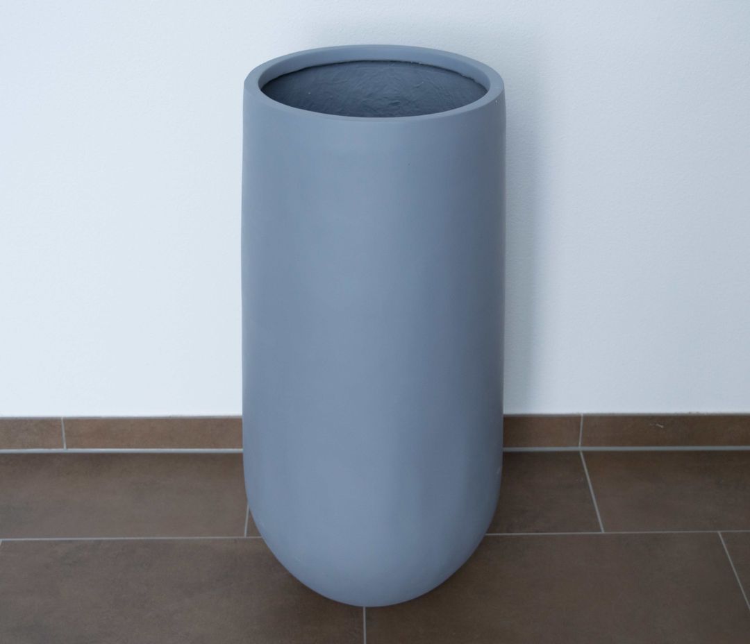 Grauer runder Pflanzkübel aus Fiberglas mit abgerundetem Boden, leer vor weißer Wand auf braunem Fliesenboden