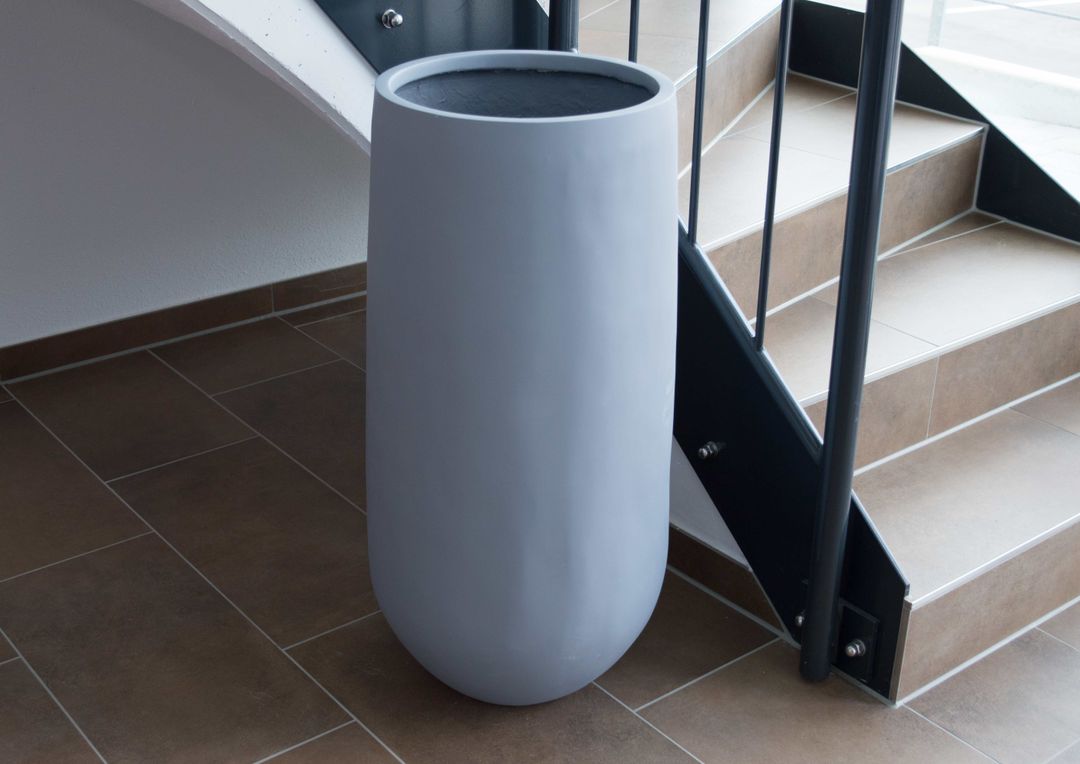 Hoher grauer Pflanzkübel aus Fiberglas in runder konischer Form, leer neben einer Treppe auf braunen Fliesen