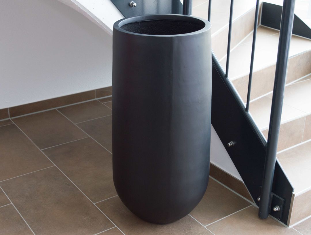 Hoher runder Pflanzkübel aus Fiberglas in Schwarz-matt, leer auf Fliesenboden neben einer Treppe mit Metallgeländer