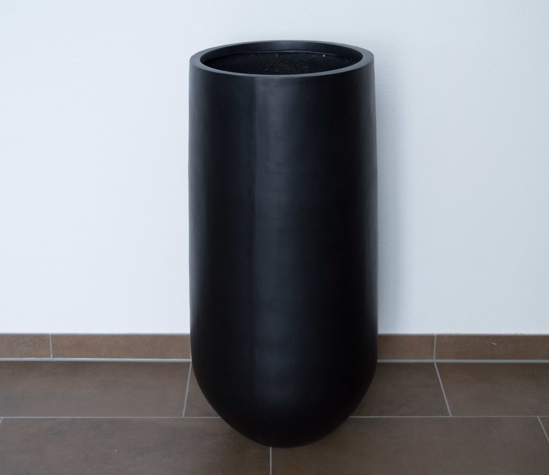 Hoher runder Pflanzkübel aus Fiberglas in Schwarz-matt, leer vor weißer Wand auf braunem Fliesenboden