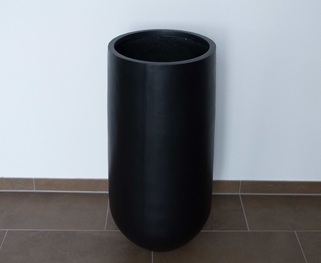 Schwarz-matter runder Pflanzkübel aus Fiberglas mit abgerundetem Boden vor weißer Wand auf braunem Fliesenboden
