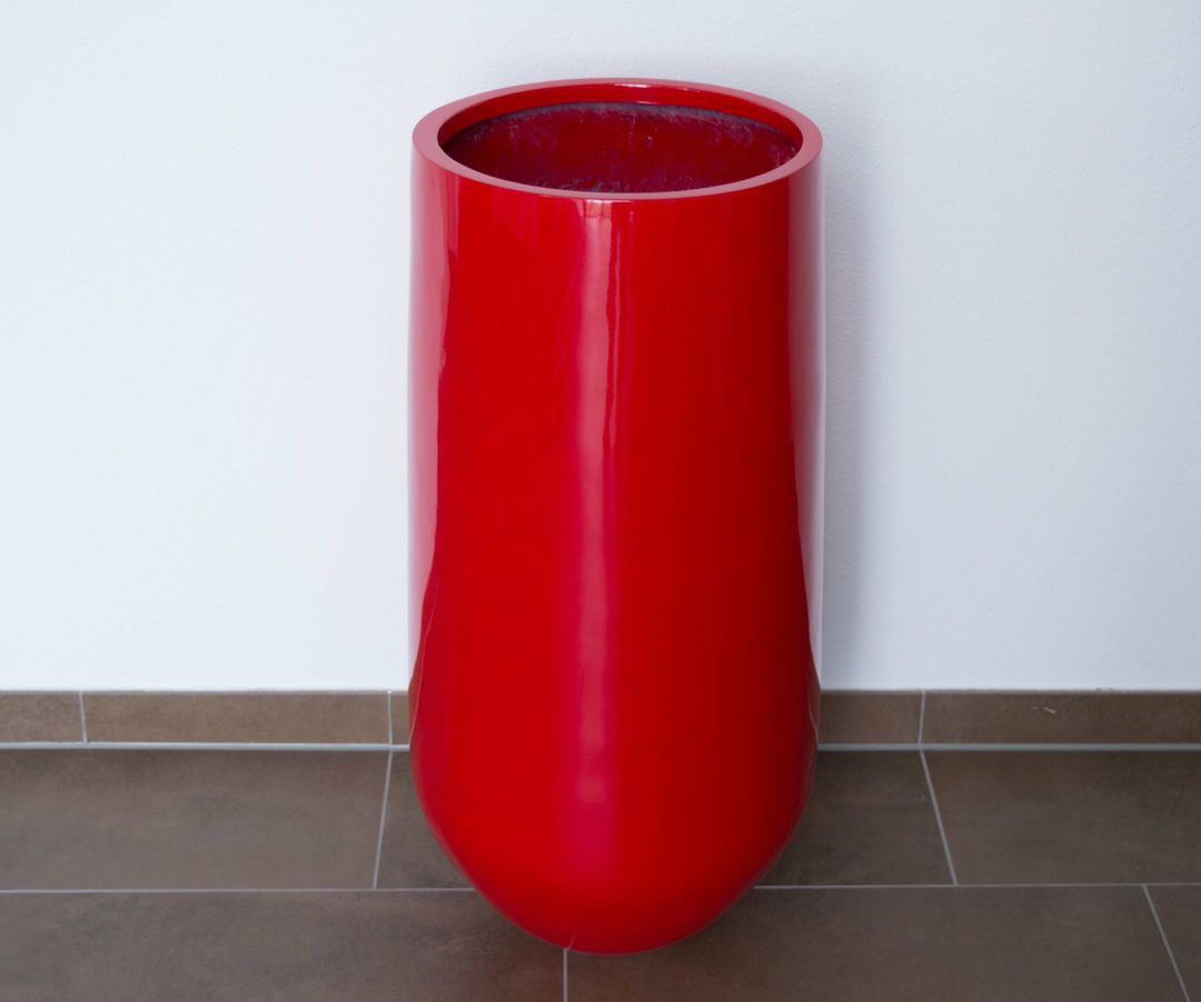 Hochglanz roter, runder konischer Pflanzkübel aus Fiberglas ohne Bepflanzung vor weißer Wand auf braunem Fliesenboden