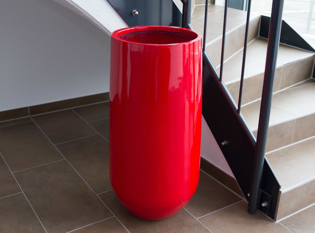 Hoher runder Pflanzkübel aus Fiberglas in Hochglanz Rot ohne Bepflanzung auf braunen Fliesen neben einer Treppe