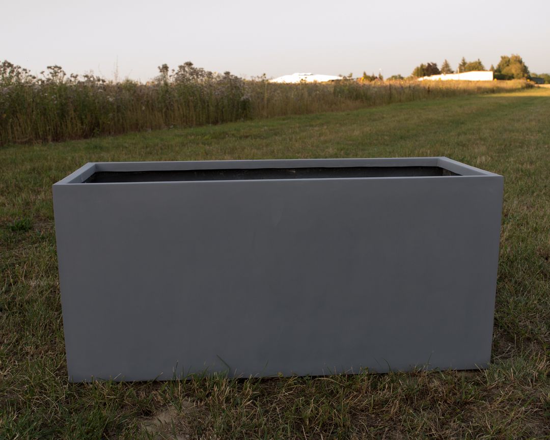 Grauer rechteckiger Pflanzkübel aus Fiberglas ohne Bepflanzung auf einer Wiese vor einem Feld im Abendlicht