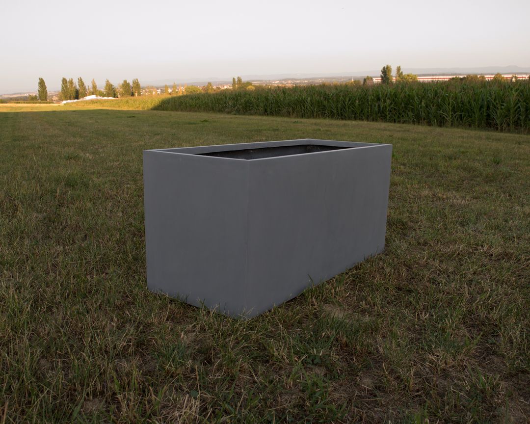Grauer rechteckiger Pflanzkübel aus Fiberglas ohne Bepflanzung auf einer Wiese vor einem Maisfeld