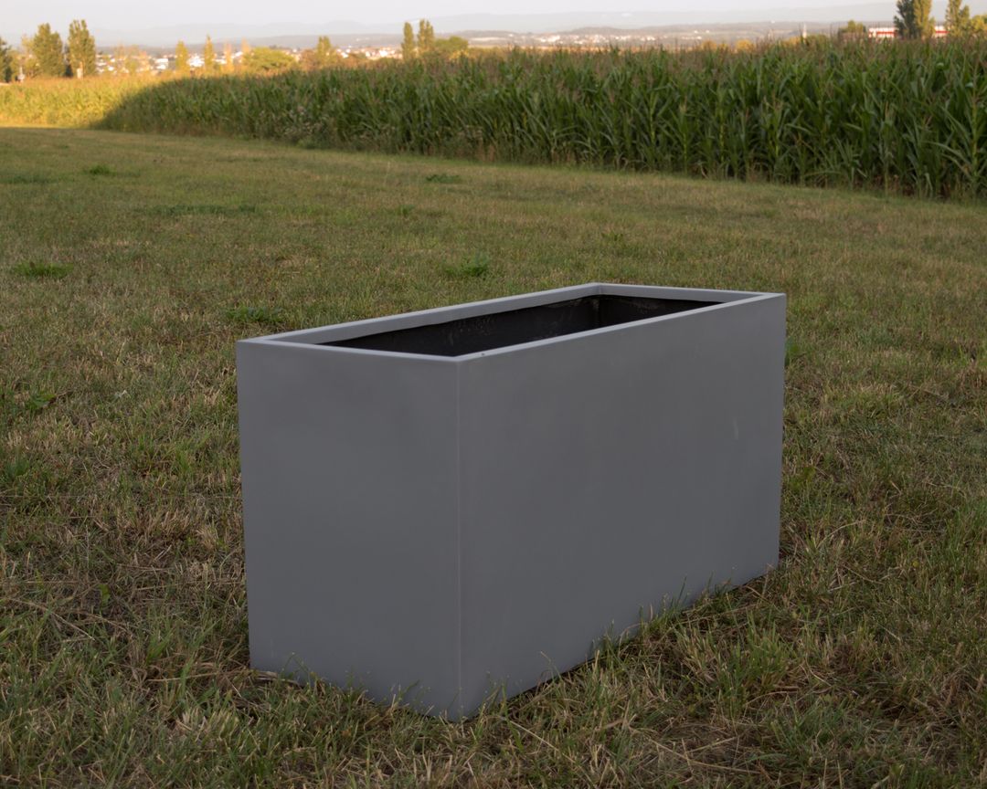 Grauer rechteckiger Pflanzkübel aus Fiberglas ohne Bepflanzung auf einer Wiese vor einem Maisfeld
