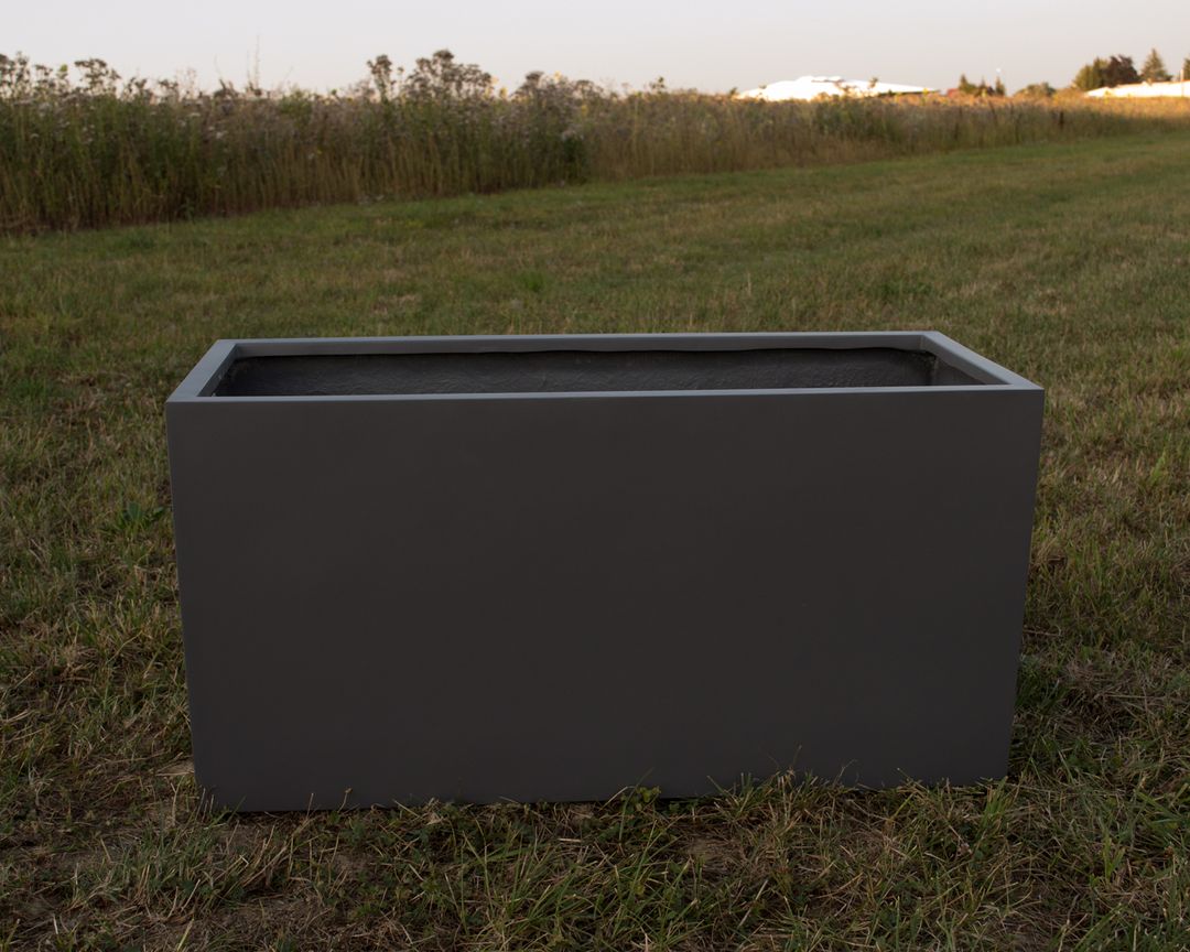 Rechteckiger Pflanzkübel aus Fiberglas in Anthrazit metallic, leer auf einer Wiese vor hohen Gräsern im Abendlicht