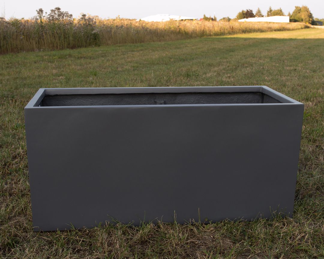 Rechteckiger Pflanzkübel aus Fiberglas in Anthrazit metallic, leer auf einer Wiese vor einem Feld im Abendlicht