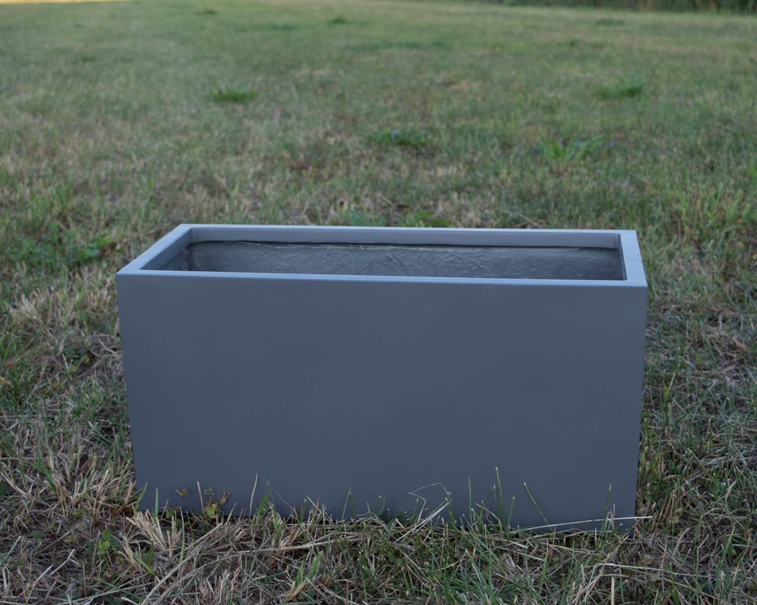 Leerer rechteckiger Pflanzkübel aus Fiberglas in Anthrazit metallic auf einer Wiese mit teils trockenem Gras