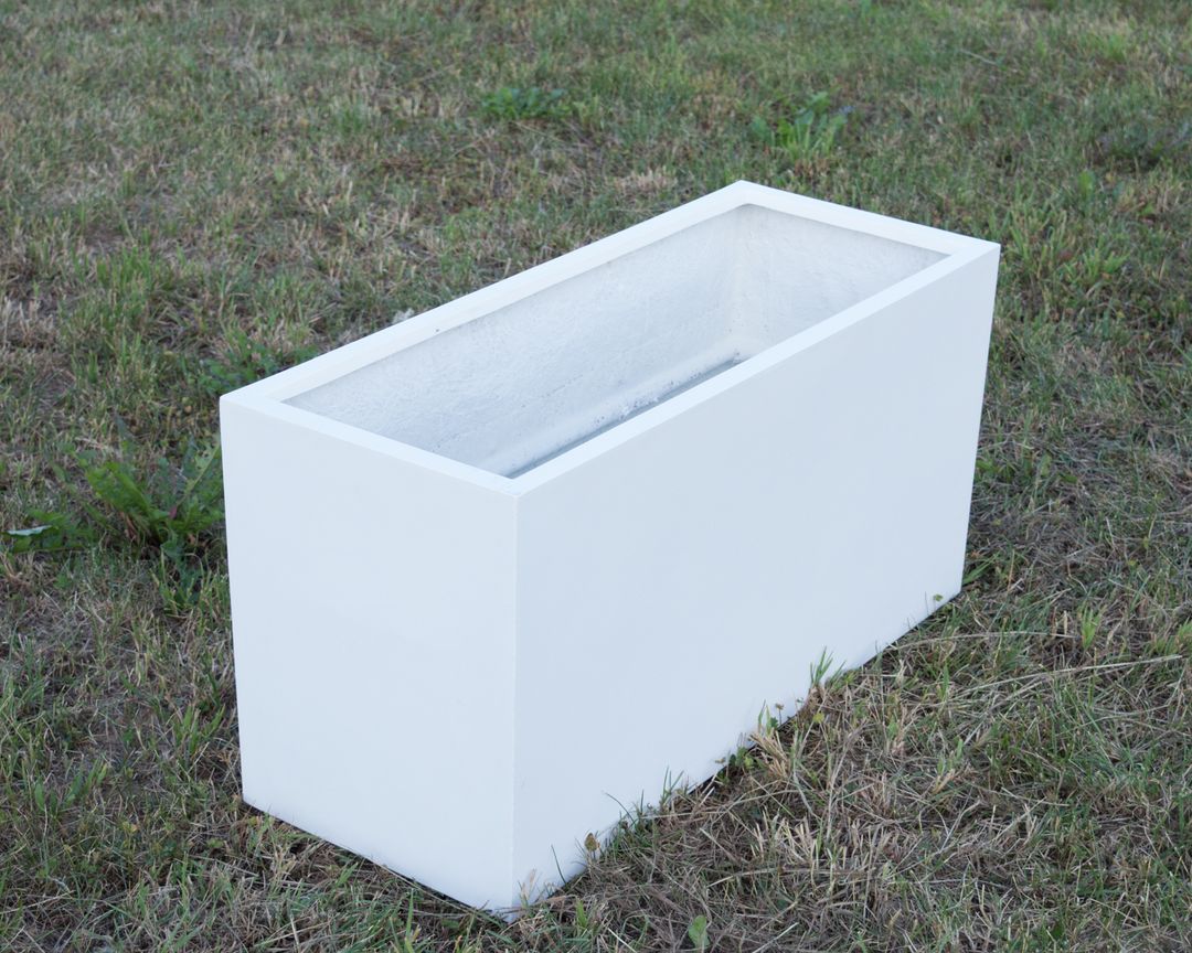 Rechteckiger Pflanzkübel aus Fiberglas in Perlmuttweiß, leer auf einer Rasenfläche, schräg von oben gesehen