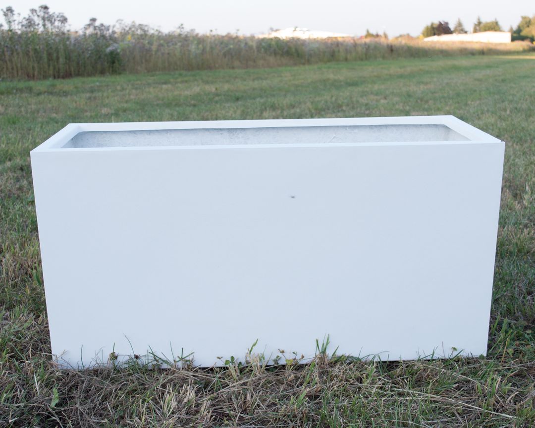 Leerer rechteckiger Pflanzkübel aus Fiberglas in Perlmuttweiß auf einer Wiese vor hohem Gras