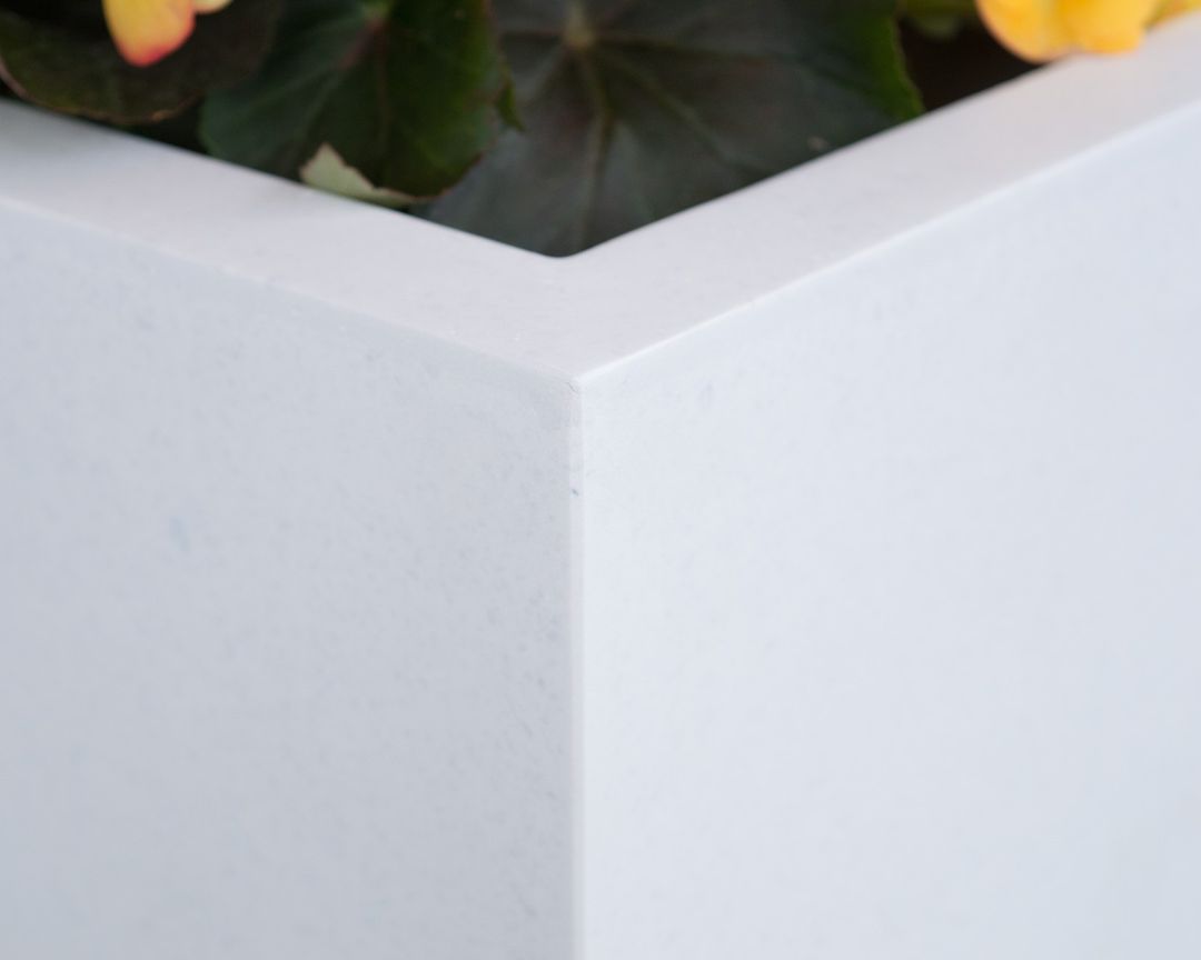 Nahaufnahme der Ecke eines perlmuttweißen rechteckigen Pflanzkübels aus Fiberglas mit gelb blühender Begonie