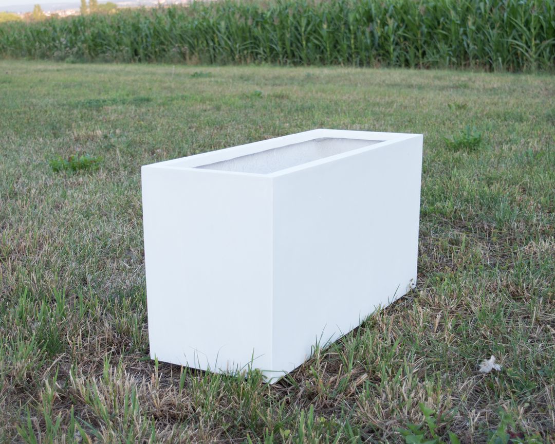 Rechteckiger Pflanzkübel aus Fiberglas in Perlmuttweiß ohne Bepflanzung auf einer Wiese vor einem Maisfeld