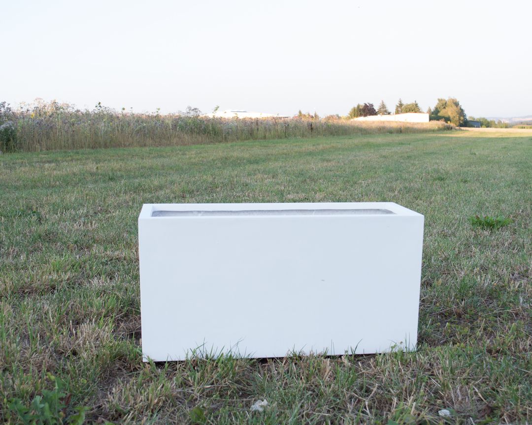 Rechteckiger Pflanzkübel aus Fiberglas in Perlmuttweiß ohne Bepflanzung auf einer Wiese vor Feldern im Abendlicht