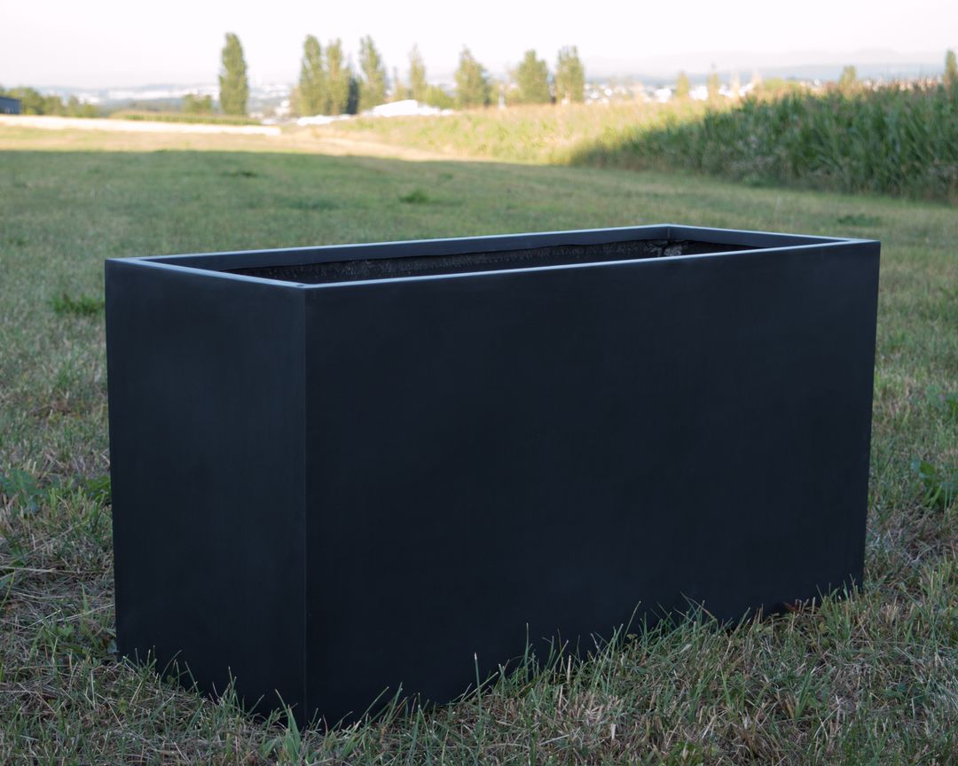 Rechteckiger Pflanzkübel aus Fiberglas in Schwarz-Matt, leer auf einer Wiese vor Feldern und Bäumen
