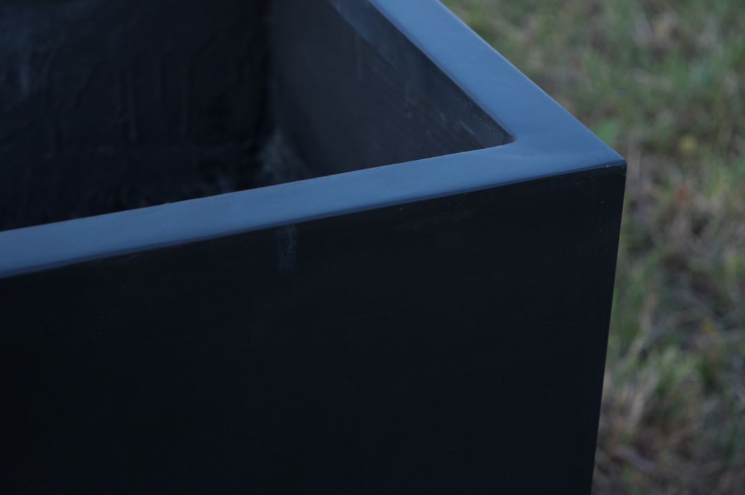 Nahaufnahme der Ecke eines rechteckigen Pflanzkübels aus Fiberglas in Schwarz-Matt mit Rand vor Rasen