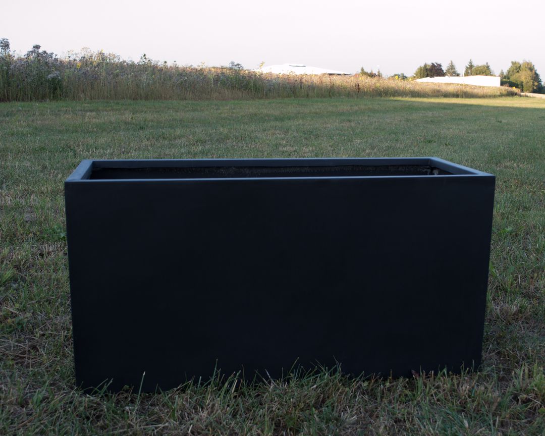 Leerer rechteckiger Pflanzkübel aus Fiberglas in Schwarz-matt auf einer Wiese vor hohem Gras und Bäumen