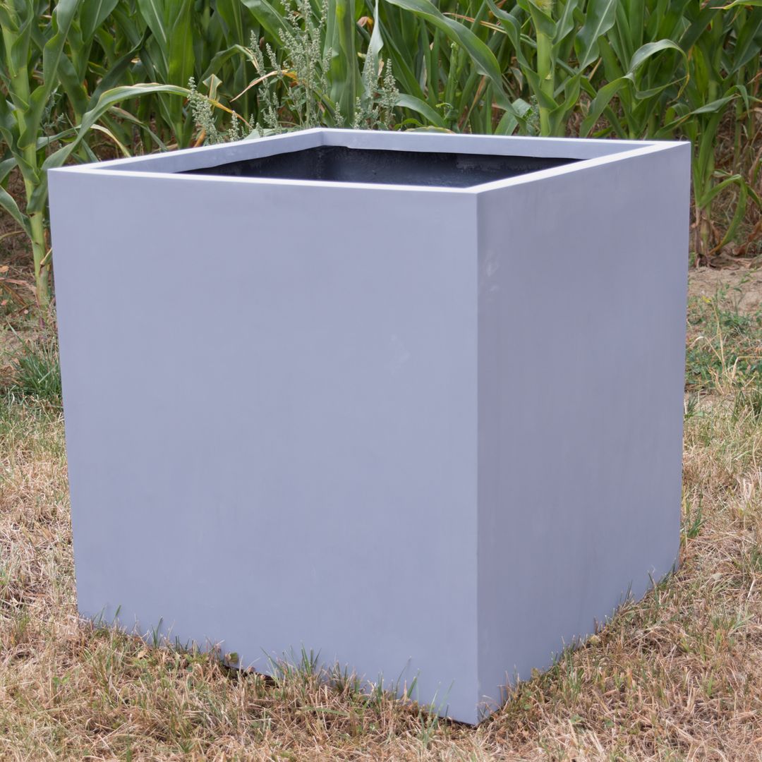 Grauer quadratischer Pflanzkübel aus Fiberglas in Würfelform auf trockenem Rasen vor einem grünen Maisfeld