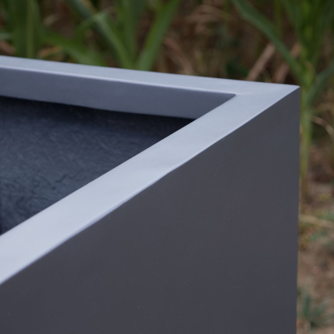 Nahaufnahme der Ecke eines quadratischen Pflanzkübels aus Fiberglas in Anthrazit metallic mit flachem Rand vor Gräsern