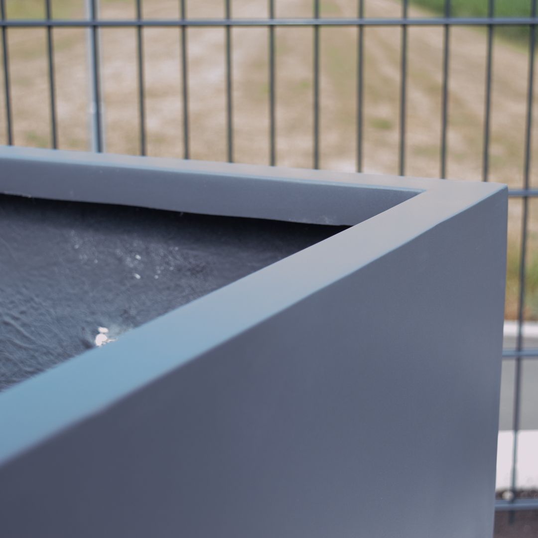 Nahaufnahme der Ecke eines quadratischen Pflanzkübels in Anthrazit metallic mit Innenrand vor einem Metallzaun