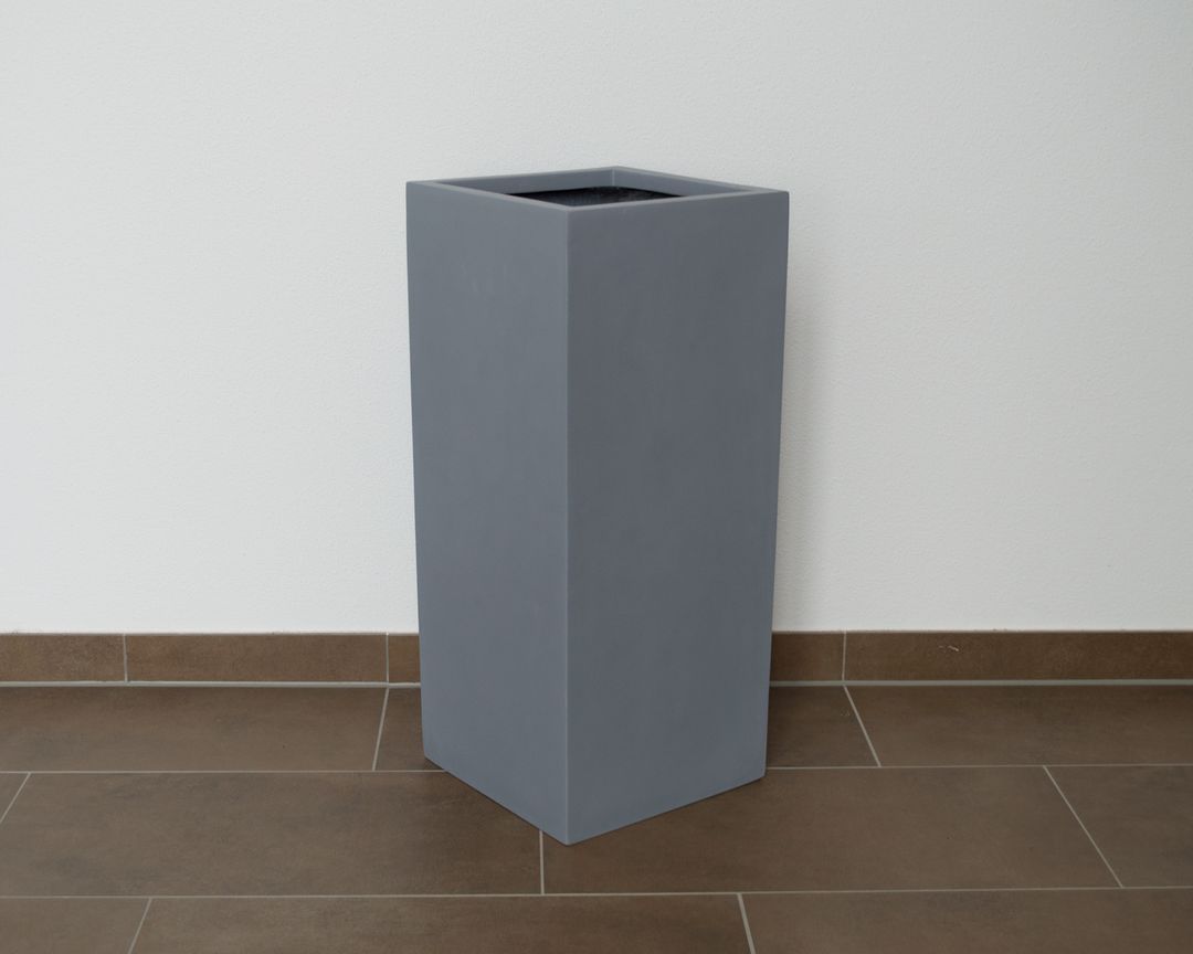 Hohe quadratische Pflanzsäule aus Fiberglas in Grau, leer vor weißer Wand auf braunem Fliesenboden