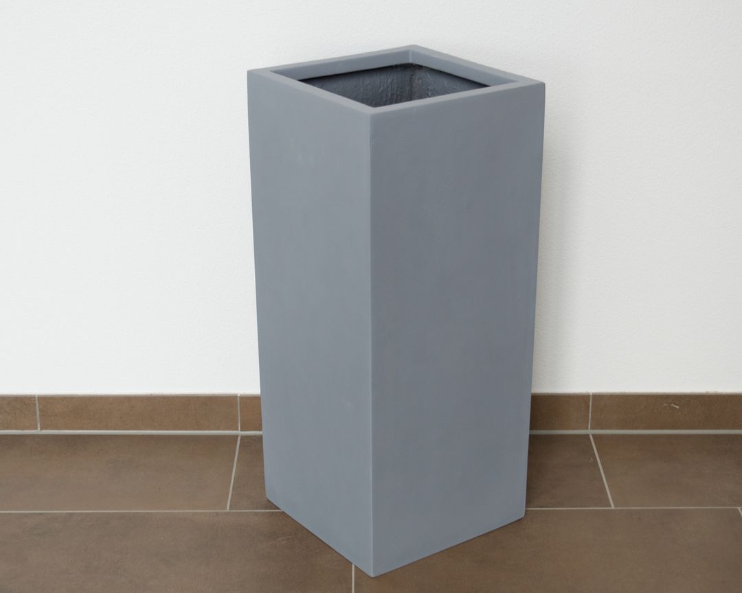 Graue quadratische Pflanzsäule aus Fiberglas ohne Bepflanzung vor weißer Wand auf braunem Fliesenboden