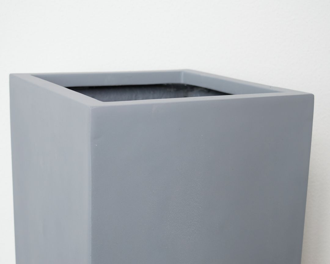Nahaufnahme des oberen Rands eines grauen, quadratischen Pflanzkübels in Säulenform aus Fiberglas vor heller Wand