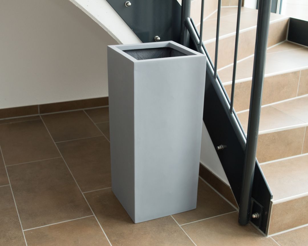Hoher quadratischer Pflanzkübel in Grau ohne Bepflanzung auf braunen Fliesen neben einer Treppe mit Metallgeländer