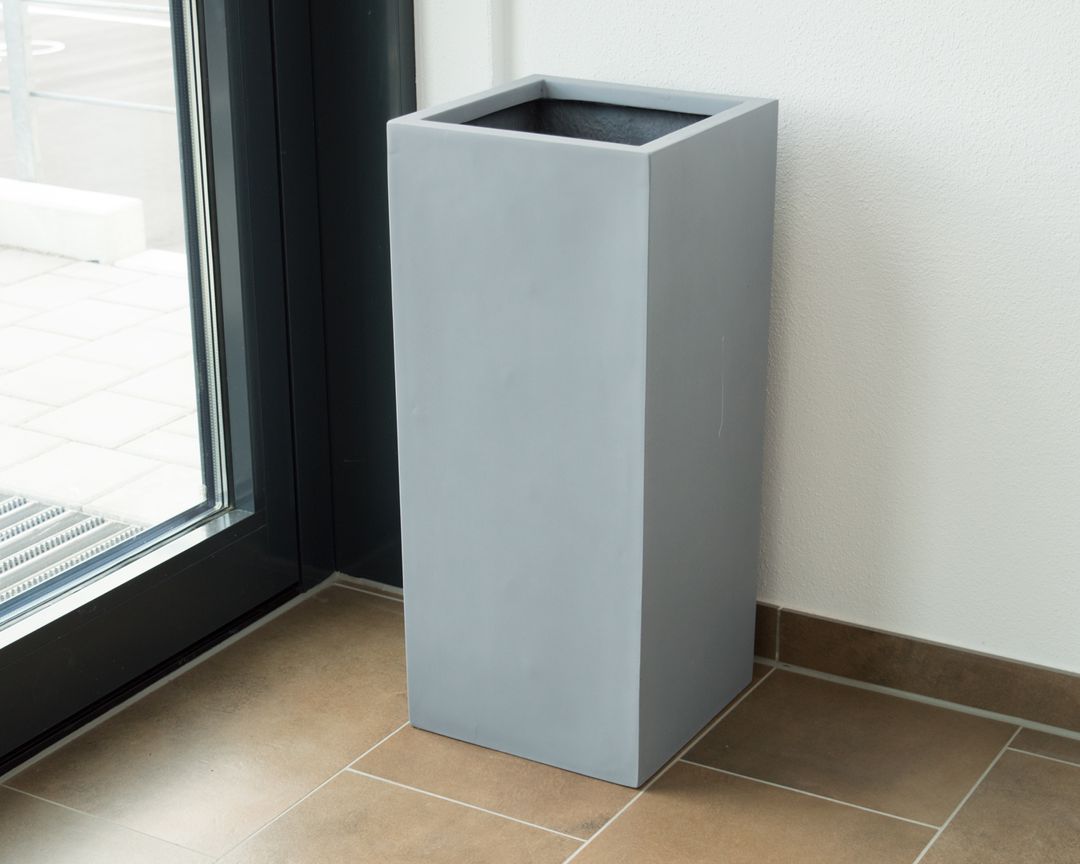 Graue quadratische Pflanzsäule aus Fiberglas ohne Bepflanzung neben einer Glastür auf braunem Fliesenboden