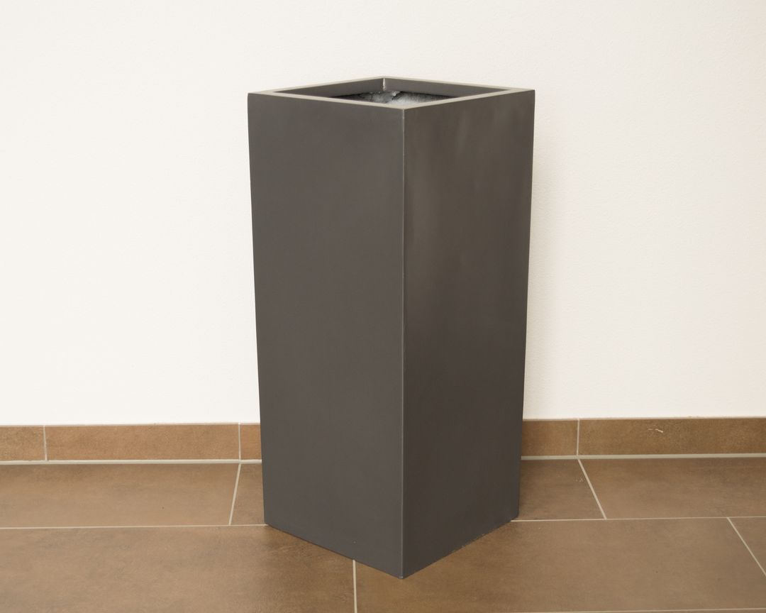 Hohe quadratische Pflanzsäule aus Fiberglas in Anthrazit metallic, unbepflanzt vor weißer Wand auf braunem Fliesenboden