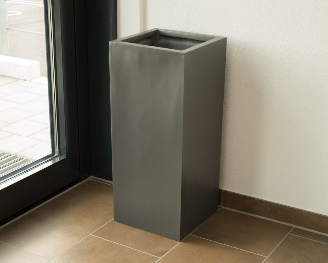 Leerer Pflanzkübel aus Fiberglas in Säulenform in Anthrazit metallic neben einer Glastür auf Fliesenboden