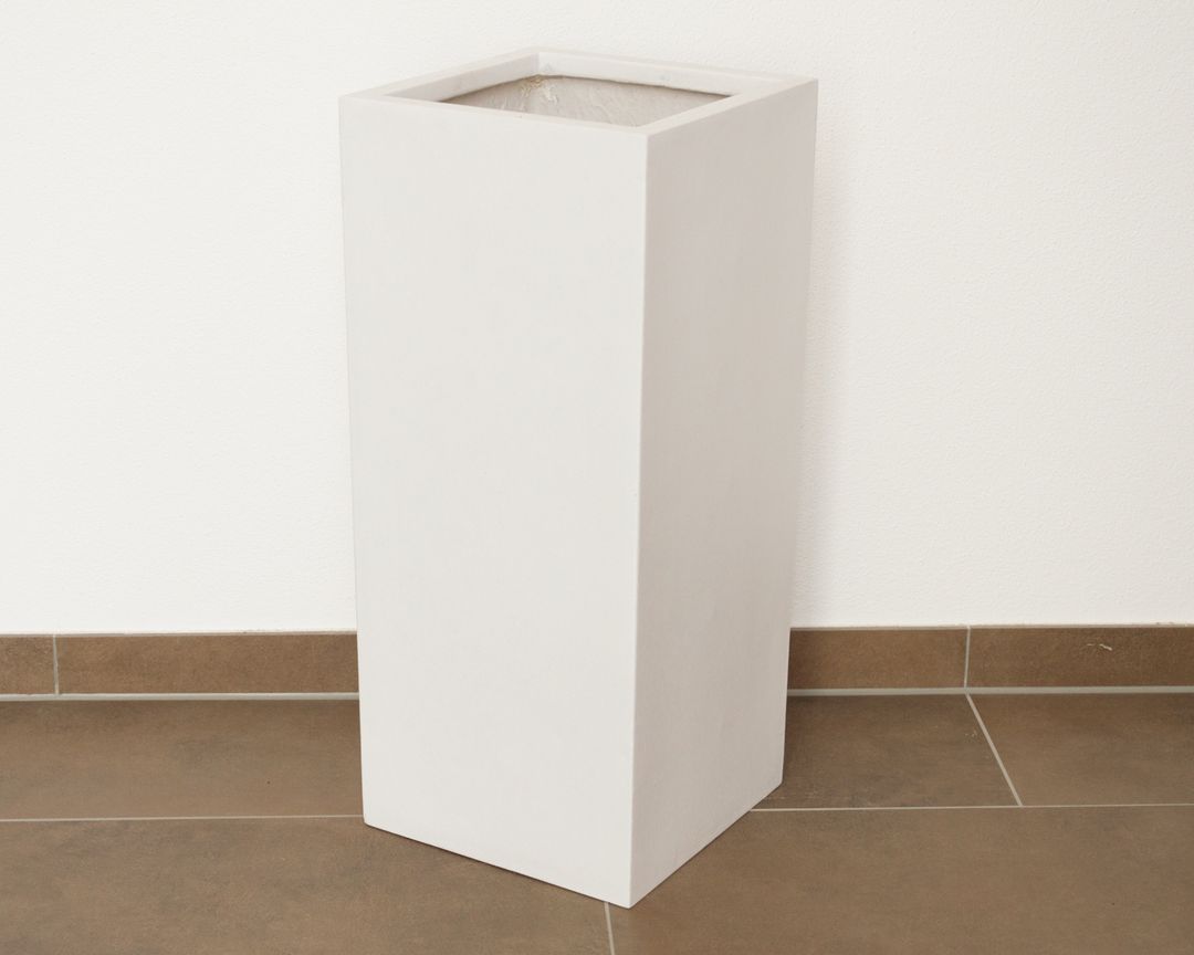 Hohe quadratische Pflanzsäule aus Fiberglas in Perlmuttweiß, leer vor weißer Wand auf braunem Fliesenboden