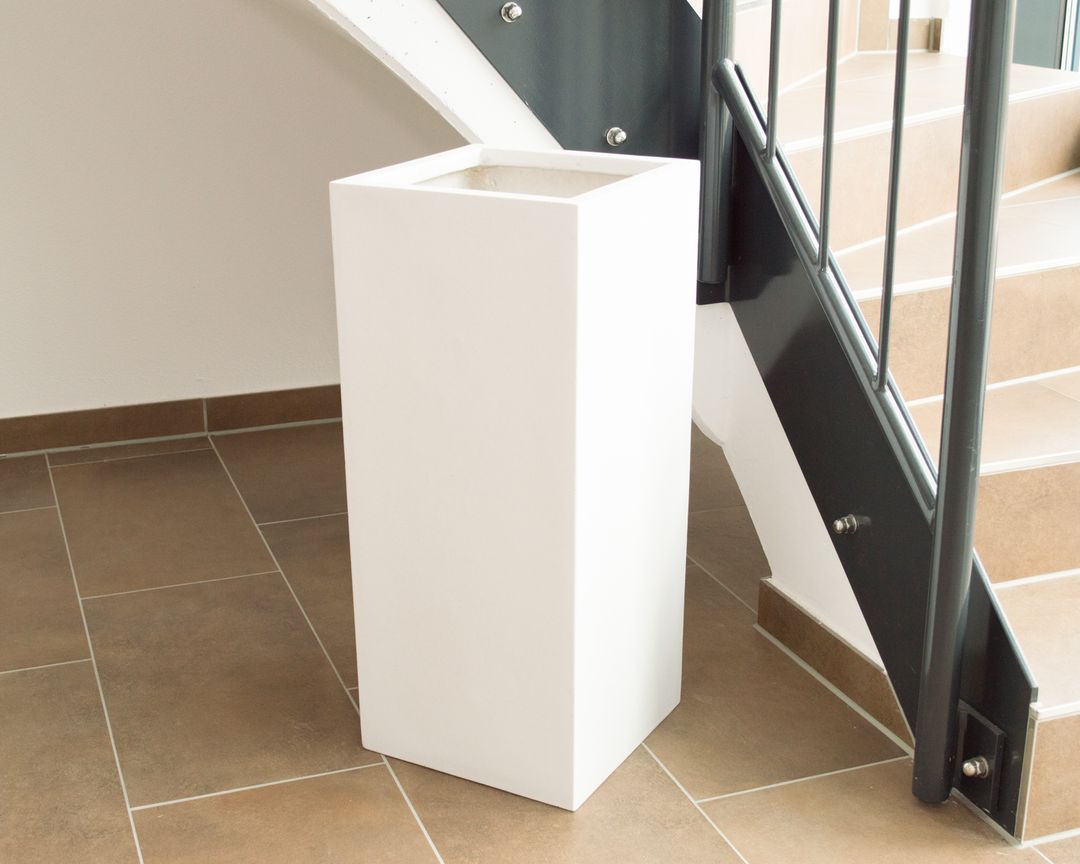 Leerer Pflanzkübel als Säule aus Fiberglas in Perlmuttweiß im Treppenhaus neben anthrazitfarbener Treppe auf Fliesenboden