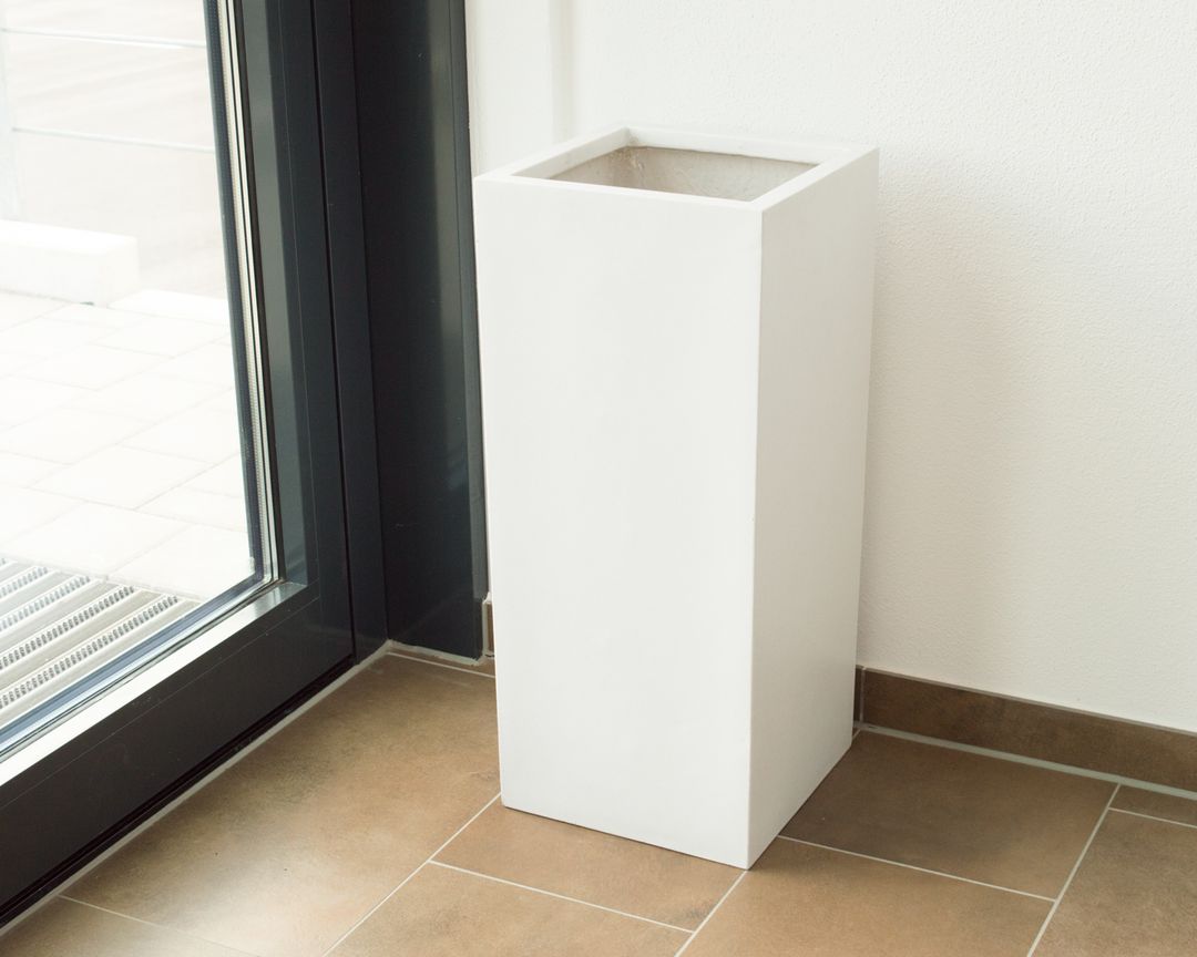 Hohe quadratische Pflanzsäule aus Fiberglas in Perlmuttweiß ohne Bepflanzung neben einer Glastür auf braunen Fliesen