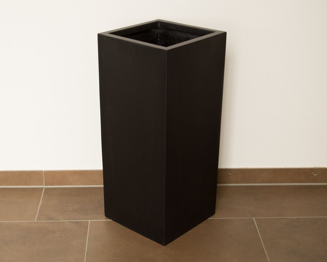 Leerer schwarz-matter Pflanzkübel aus Fiberglas in quadratischer Säulenform vor weißer Wand auf braunen Fliesen